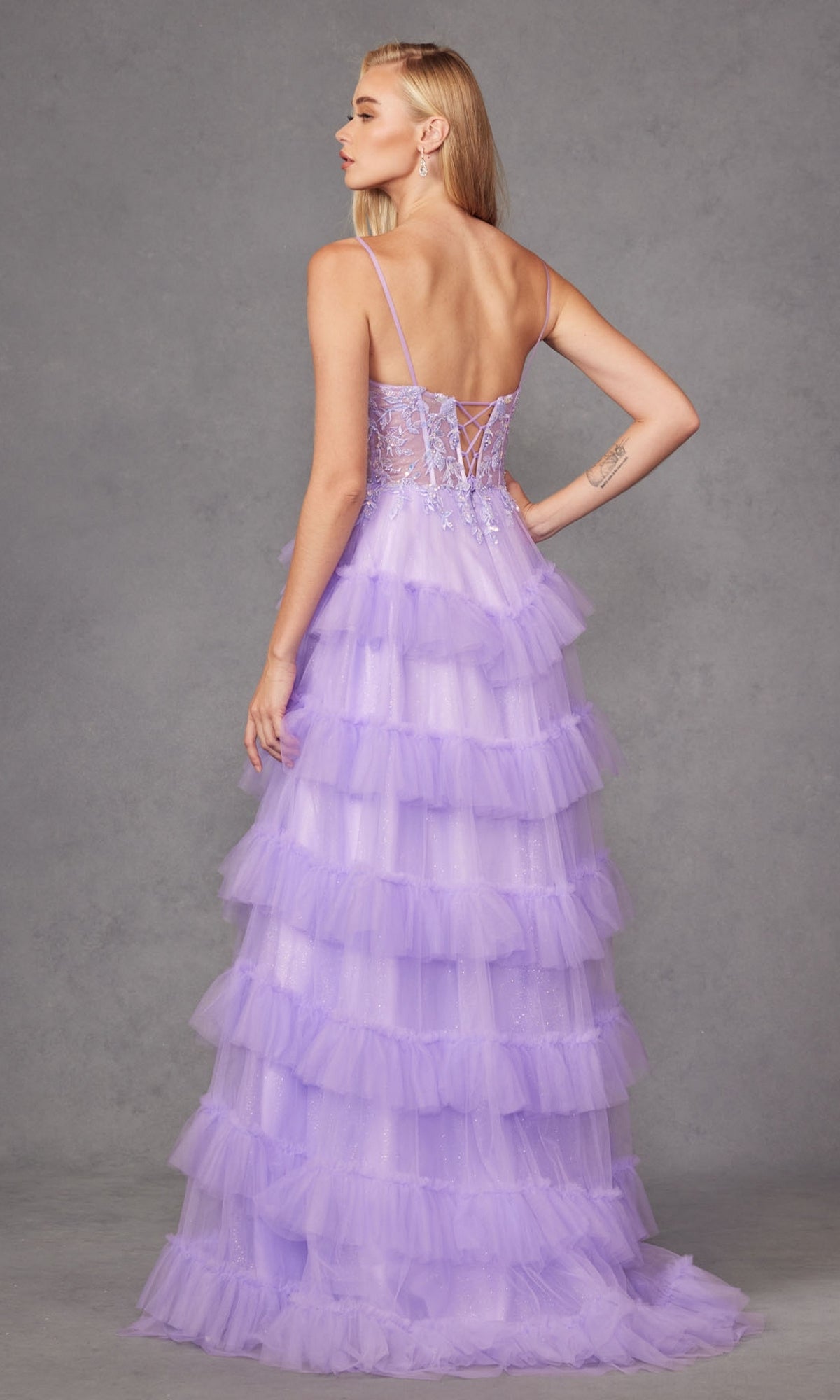 A woman faces away in the Long Prom Dress JT2459A by Juliet, a pastel purple ruffle tulle ball gown with thin straps and lace-up back, set against a plain gray background.